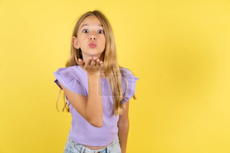 Foto de Rubia chica vistiendo violeta camiseta sobre fondo amarillo mirando a la cámara soplando un beso con la mano en el aire siendo encantador y sexy. Expresión de amor. - Imagen libre de derechos