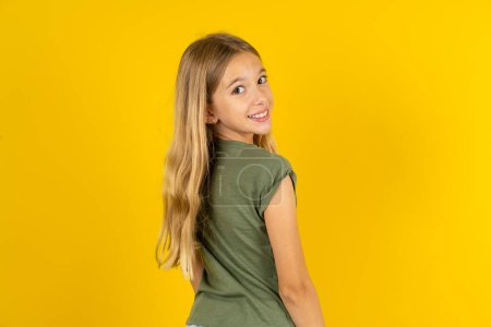 Téléchargez les photos : Satisfait jolie petite fille blonde portant chemisier kaki sur fond jaune rayonnant sourire regarder caméra - en image libre de droit