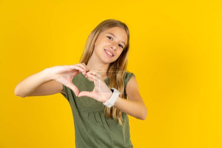 Foto de Niña rubia vistiendo blusa caqui sobre fondo amarillo sonriendo en amor haciendo forma de símbolo del corazón con las manos. Concepto romántico. - Imagen libre de derechos