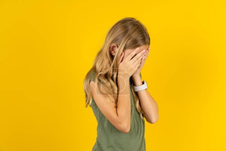 Photo for Sad blond little girl wearing khaki blouse over yellow background crying covering her face with her hands. - Royalty Free Image