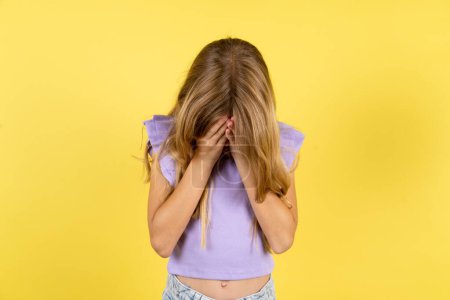 Foto de Chica rubia vistiendo camiseta violeta sobre fondo amarillo cubriendo su cara con sus manos, siendo devastado y llorando. Triste concepto - Imagen libre de derechos