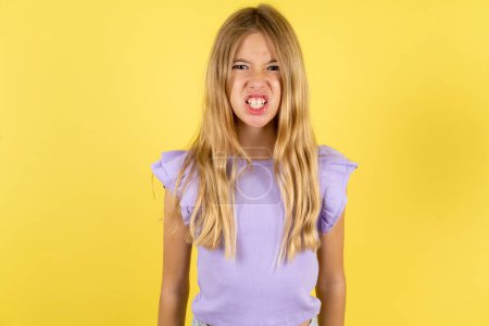 Photo for Blonde girl wearing violet T-shirt over yellow background keeps teeth clenched, frowns face in dissatisfaction, irritated because of much duties. - Royalty Free Image