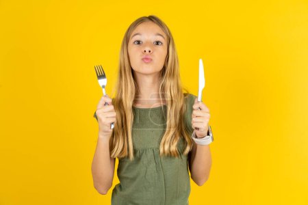 Photo for Hungry blond little girl wearing khaki blouse over yellow background holding in hand fork knife want tasty yummy pizza pie - Royalty Free Image