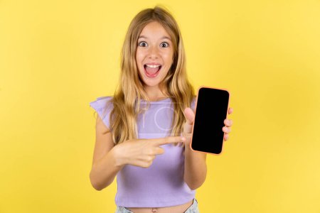 Téléchargez les photos : Attrayant gai blonde fille portant violet T-shirt sur fond jaune tenant dans les mains cellule montrant écran noir - en image libre de droit