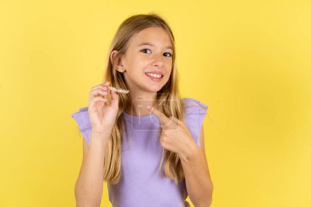 Téléchargez les photos : Fille blonde portant violet T-shirt sur fond jaune tenant un aligneur invisible et pointant vers elle. Concept de soins dentaires et de confiance. - en image libre de droit