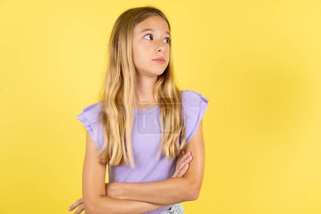 Photo for Charming thoughtful blonde girl wearing violet T-shirt over yellow background stands with arms folded concentrated somewhere with pensive expression thinks what to do - Royalty Free Image