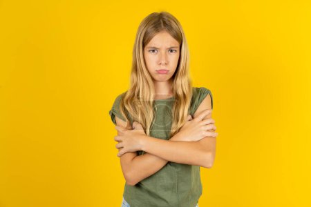 Photo for Gloomy dissatisfied blond little girl wearing khaki blouse over yellow background looks with miserable expression at camera from under forehead, makes unhappy grimace - Royalty Free Image