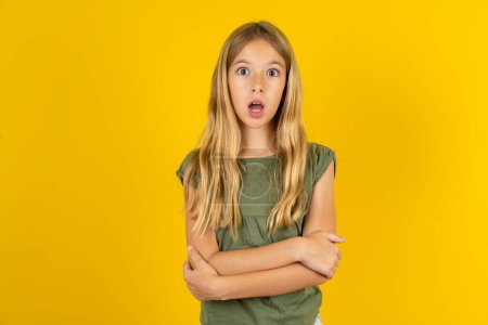 Photo for Shocked embarrassed blond little girl wearing khaki blouse over yellow background keeps mouth widely opened. Hears unbelievable novelty stares in stupor - Royalty Free Image