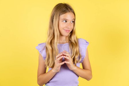 Photo for Blonde girl wearing violet T-shirt over yellow background steepled fingers and looks mysterious aside has great evil plan in mind - Royalty Free Image