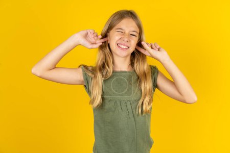 Téléchargez les photos : Joyeux blond petite fille portant chemisier kaki sur fond jaune ignore la musique forte et bouchons les oreilles avec les doigts demande d'éteindre le son - en image libre de droit