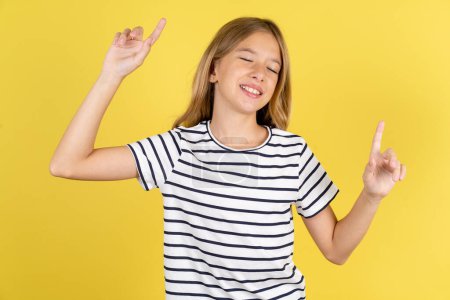 Téléchargez les photos : Photo de belle adolescente caucasienne optimiste portant T-shirt rayé sur fond jaune a du plaisir et danse vêtements insouciants être d'humeur parfaite fait des mouvements. Passe du temps libre sur disco partie - en image libre de droit