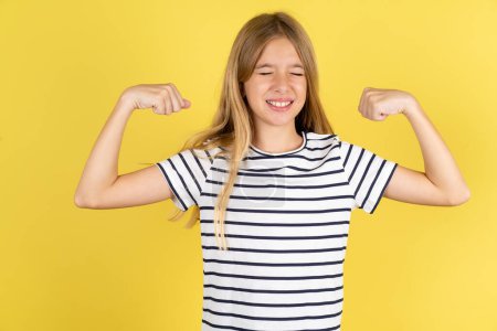 Photo for Strong powerful beautiful caucasian teen girl wearing striped T-shirt over yellow background toothy smile, raises arms and shows biceps. Look at my muscles! - Royalty Free Image