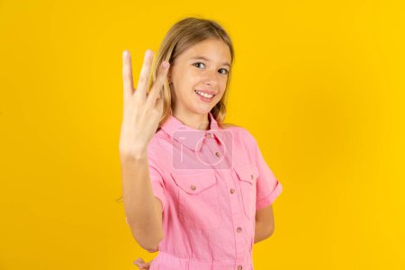 Foto de Niña vistiendo chaqueta rosa sobre fondo amarillo sonriendo y mirando amigable, mostrando el número tres o tercero con la mano hacia adelante, cuenta atrás - Imagen libre de derechos