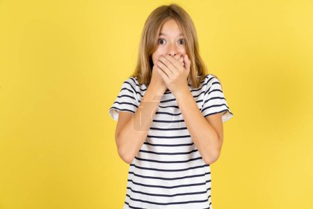 Foto de Aturdido hermosa chica adolescente caucásica con rayas camiseta sobre fondo amarillo cubre ambas manos en la boca, temores de algo asombroso - Imagen libre de derechos