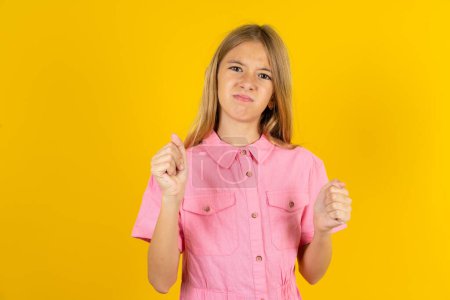 Photo for Irritated girl wearing pink jacket over yellow background blows cheeks with anger and raises clenched fists expresses rage and aggressive emotions. Furious model - Royalty Free Image
