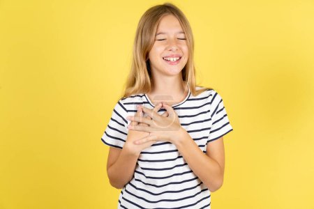 Téléchargez les photos : Belle adolescente caucasienne portant T-shirt rayé sur fond jaune exprime le bonheur, rit agréablement, garde les mains sur le cœur - en image libre de droit