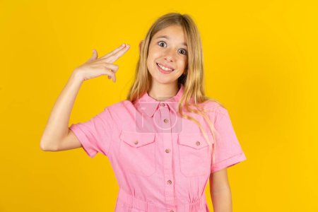 Foto de Chica vistiendo chaqueta rosa sobre fondo amarillo tontería alrededor de los brotes en la sien con los dedos hace gesto suicida. Modelo divertido hace pistola de dedo - Imagen libre de derechos