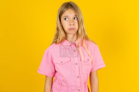 Photo for Dissatisfied girl wearing pink jacket over yellow background purses lips and has unhappy expression looks away stands offended. Depressed frustrated model. - Royalty Free Image