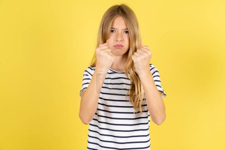 Téléchargez les photos : Displeased annoyed beautiful caucasian teen girl wearing striped T-shirt over yellow background clenches fists, gestures pissed, ready to revenge, looks with aggression at camera stands full of hate, being pressured - en image libre de droit