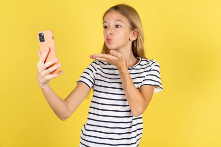Téléchargez les photos : Belle fille adolescente caucasienne portant T-shirt rayé sur fond jaune souffle baiser d'air à la caméra du smartphone et prend selfie, envoie mwah via appel en ligne. - en image libre de droit