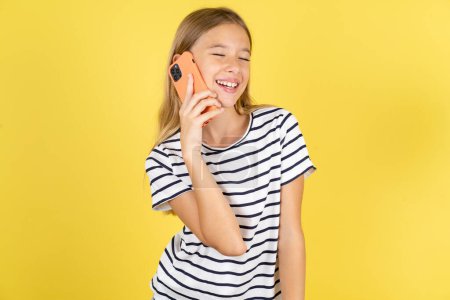 Foto de Divertido hermosa chica adolescente caucásica con rayas camiseta sobre fondo amarillo ríe felizmente, tiene conversación telefónica, siendo divertido por amigo, cierra los ojos. - Imagen libre de derechos