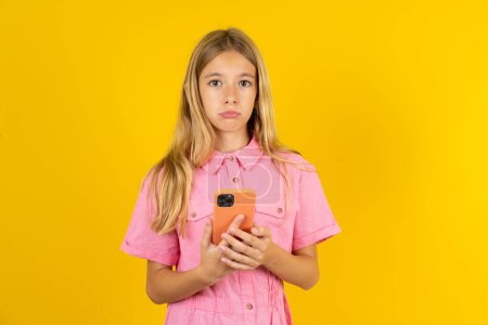 Foto de Niña molesta insatisfecha con chaqueta rosa sobre fondo amarillo utiliza la aplicación de software móvil y navega información en Internet, sostiene la mano móvil moderna - Imagen libre de derechos