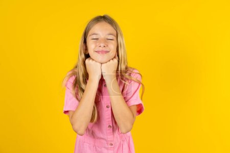 Foto de Chica alegre con chaqueta rosa sobre fondo amarillo tiene expresión satisfecha tímida, sonríe ampliamente, muestra dientes blancos, emociones de la gente - Imagen libre de derechos