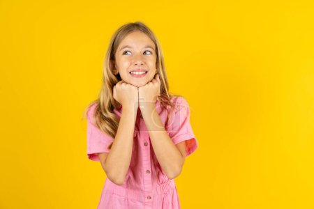 Téléchargez les photos : Fille portant une veste rose sur fond jaune tient les mains sous le menton, heureux d'entendre des mots chaleureux de l'étranger - en image libre de droit