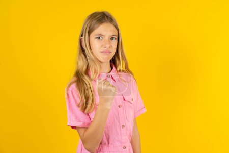 Photo for Girl wearing pink jacket over yellow background shows fist has annoyed face expression going to revenge or threaten someone makes serious look. I will show you who is boss - Royalty Free Image