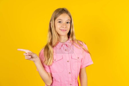 Photo for Girl wearing pink jacket over yellow background points to side on blank space demonstrates advertisement. People and promotion concept - Royalty Free Image