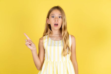 Photo for Emotive girl wearing yellow dress over yellow background keeps jaw dropped from shock demonstrates amazing promo points right on blank space demonstrates big shopping sale. Advertisement - Royalty Free Image
