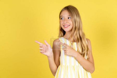 Téléchargez les photos : Fille portant robe jaune sur fond jaune sourire excité doigts directifs regarder espace vide - en image libre de droit