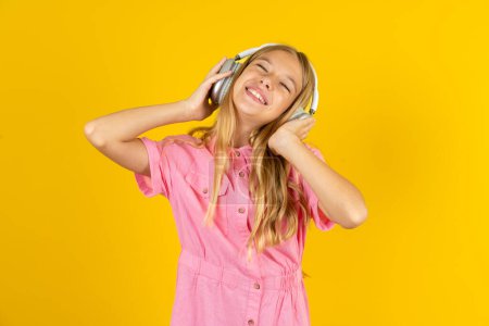 Foto de Chica con chaqueta rosa sobre fondo amarillo sonrisas ampliamente se siente muy contento escucha pista de música favorita a través de auriculares inalámbricos cierra los ojos. - Imagen libre de derechos