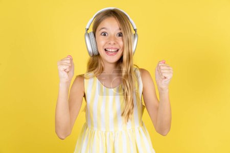Photo for Emotional girl wearing yellow dress over yellow background exclaims loudly feels like winner raises clenched fists keeps mouth opened wears stereo headphones on ears makes yes gesture, listens favorite music - Royalty Free Image