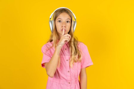 Foto de Niña con chaqueta rosa sobre fondo amarillo haciendo gesto de silencio con el dedo en los labios usando auriculares inalámbricos. Silencio.. - Imagen libre de derechos