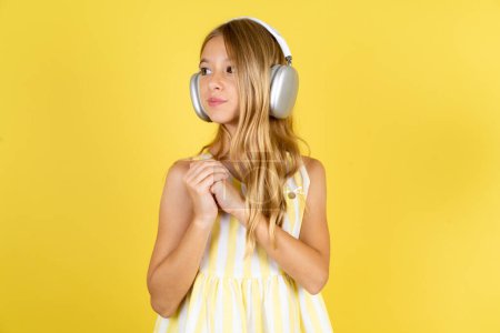 Téléchargez les photos : Fille portant une robe jaune sur fond jaune porte un casque stéréo écouter de la musique concentrée et regarder de côté avec intérêt. - en image libre de droit