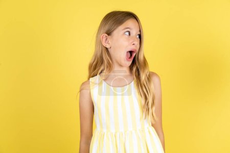 Schockiertes Mädchen in gelbem Kleid vor gelbem Hintergrund sieht mit offenem Mund leer aus und schreit: Oh mein Gott! Ich kann das nicht glauben.