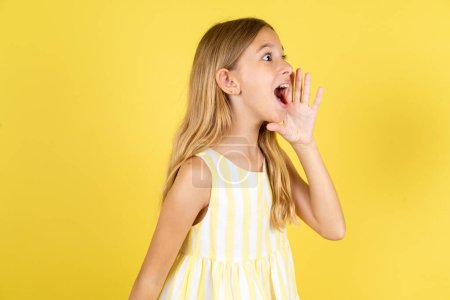 Téléchargez les photos : Fille portant une robe jaune sur fond jaune regarder espace vide tenant le visage de la main et crier ou appeler quelqu'un. - en image libre de droit