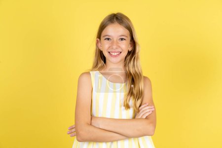 Téléchargez les photos : Portrait de charmante fille portant une robe jaune sur fond jaune debout avec confiance souriant avec les mains pliées - en image libre de droit