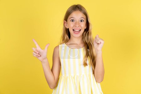 Téléchargez les photos : Fille portant une robe jaune sur fond jaune points à l'espace vide tenant le poing vers le haut, geste gagnant. - en image libre de droit
