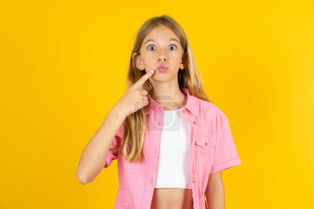 Photo for Charming girl wearing pink jacket over yellow background pointing on pout lips with forefinger, showing effect after lifting procedure, - Royalty Free Image