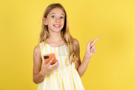 Foto de Chica asombrada vistiendo vestido amarillo sobre fondo amarillo sosteniendo su teléfono y señalando con el dedo a un lado en el espacio de copia vacío - Imagen libre de derechos
