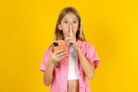Téléchargez les photos : Fille portant une veste rose sur fond jaune tenant gadget moderne demander de ne pas dire de secrets - en image libre de droit