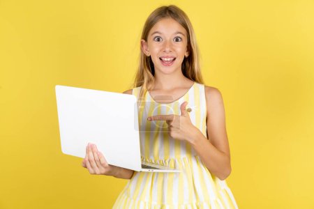 Photo for Shocked girl wearing yellow dress over yellow background pointing finger modern device - Royalty Free Image