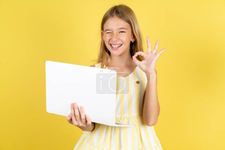 Téléchargez les photos : Attrayant gai fille habillée portant robe jaune sur fond jaune à l'aide d'un ordinateur portable montrant ok-signe winkin - en image libre de droit