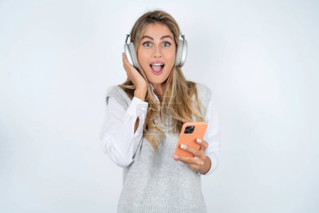 Téléchargez les photos : Belle femme d'affaires blonde hispanique positive sur fond blanc tient un téléphone portable moderne connecté à des écouteurs, serre le poing de bonnes émotions, s'exclame avec joie, - en image libre de droit
