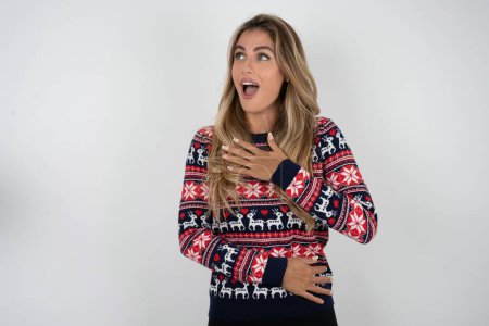 Photo for Joyful beautiful blonde woman wearing knitted christmas sweater expresses positive emotions recalls something funny keeps hand on chest and giggles happily. - Royalty Free Image