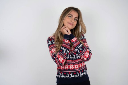 Téléchargez les photos : Charme grave Belle femme blonde portant un pull de Noël tricoté garde les mains près du visage sourit tendrement à la caméra - en image libre de droit