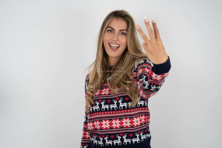 Photo for Beautiful blonde woman wearing knitted christmas sweater smiling and looking friendly, showing number three or third with hand forward, counting down - Royalty Free Image