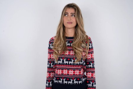Photo for Dissatisfied beautiful blonde woman wearing knitted christmas sweater purses lips and has unhappy expression looks away stands offended. Depressed frustrated model. - Royalty Free Image